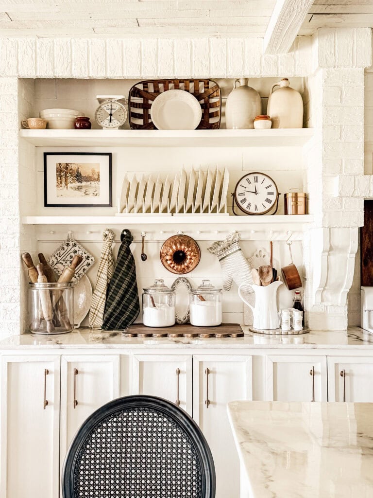 A bright, cozy kitchen with white cabinets, open shelves holding dishes, jars, and decor, a black chair in the foreground, and a small stove topped with glass canisters and a white pitcher.