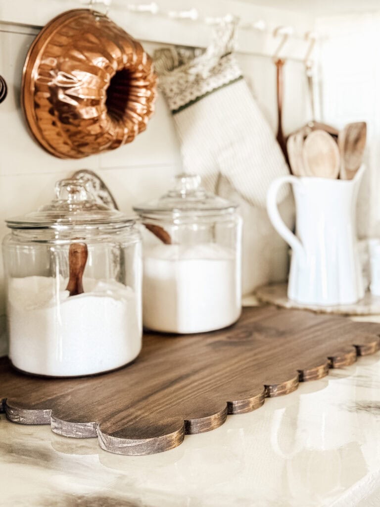 Two glass jars filled with sugar and flour, each with a wooden scoop, sit on a wooden tray in a bright kitchen. A copper bundt pan, oven mitt, and wooden utensils in a white pitcher are in the background.