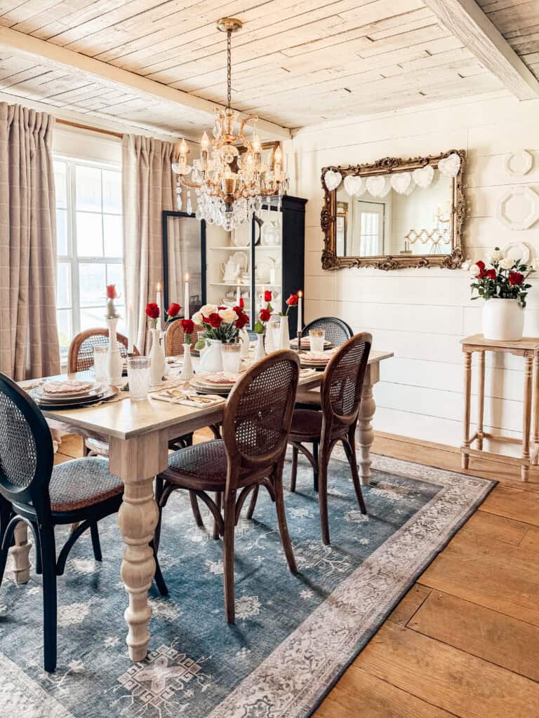 A cozy dining room with a rustic wooden table set for six, wicker chairs, a chandelier, fresh red roses in vases, a large ornate mirror, light curtains, and a blue patterned rug on wooden floors.