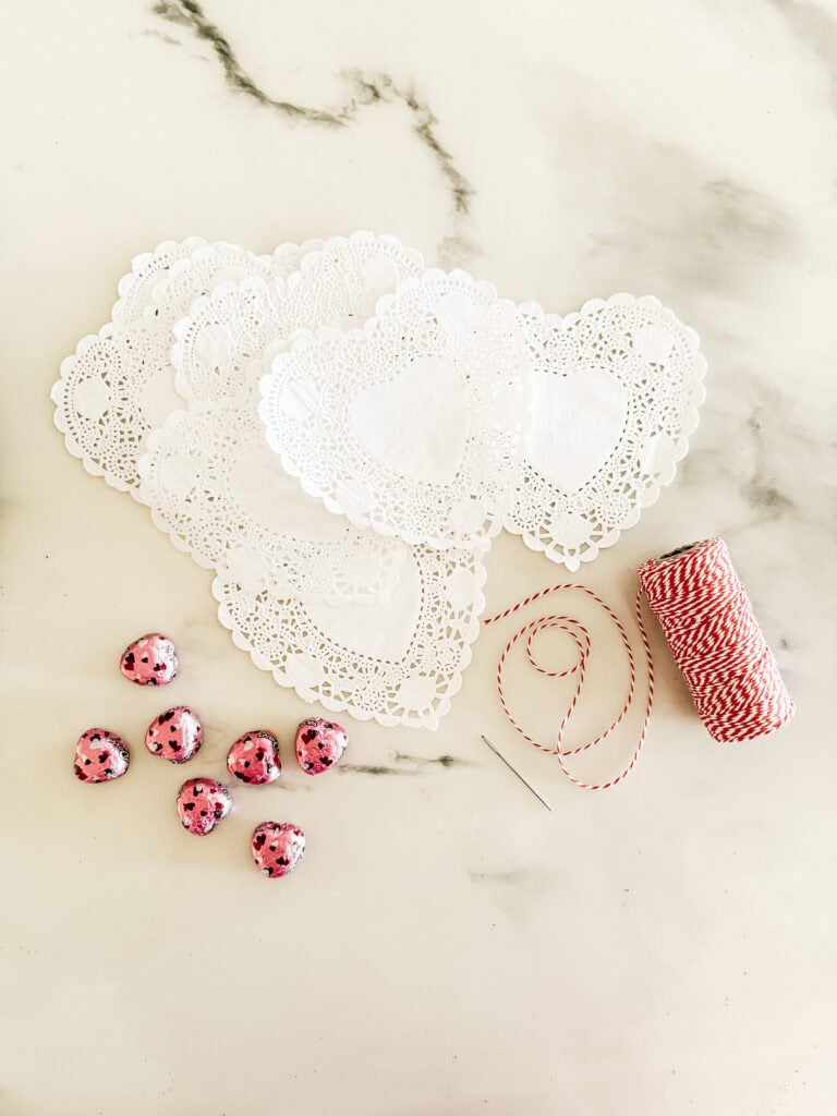 Heart-shaped paper doilies, red and white baker’s twine, a needle, and foil-wrapped heart chocolates are arranged on a white marble surface.