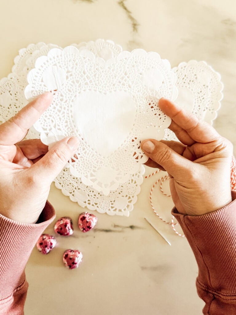 A person holds a heart-shaped white paper doily over a matching stack on a table, with pink foil-wrapped heart chocolates, red and white twine, and a sewing needle nearby.