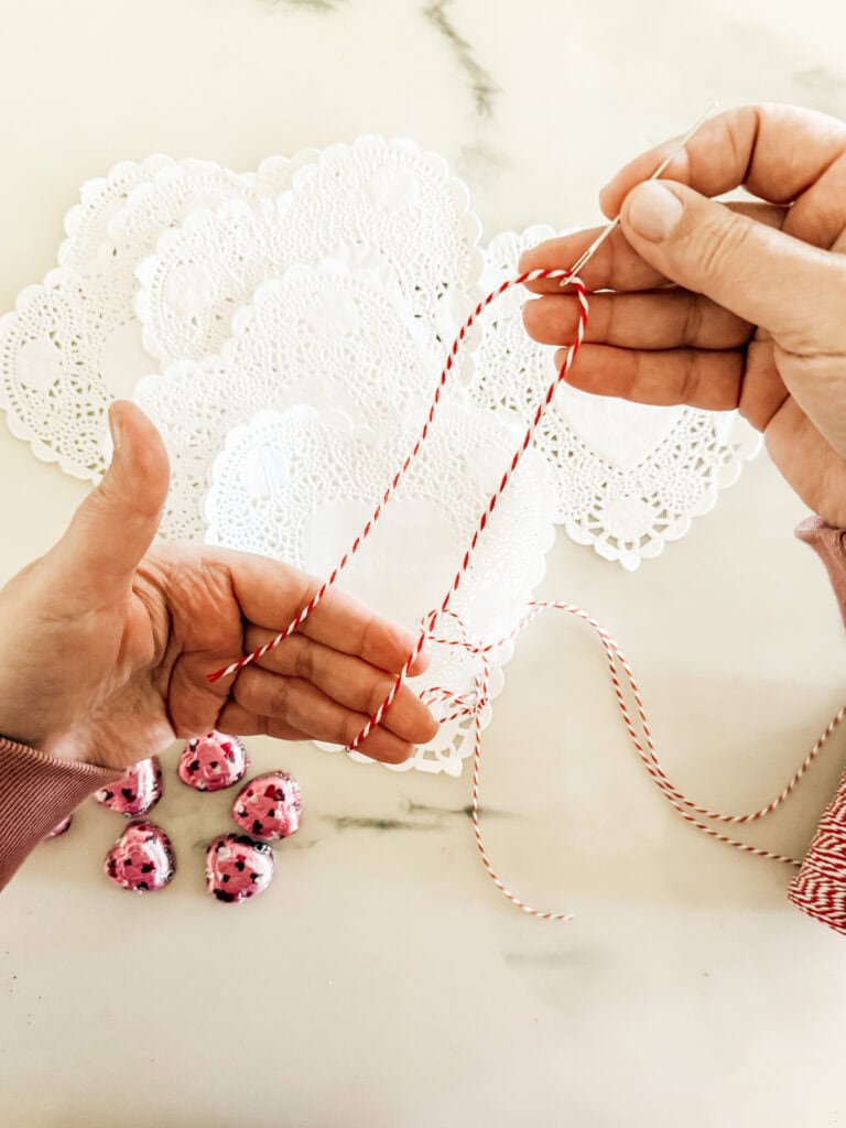 Hands threading red and white twine through a needle, with white paper doilies and pink foil-wrapped chocolate hearts on a marble surface in the background.