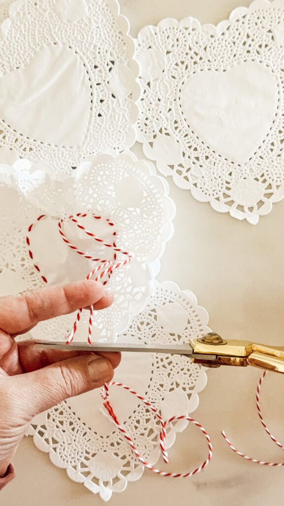A hand holds gold scissors, cutting red and white twine next to several white paper doilies with heart patterns on a light surface.