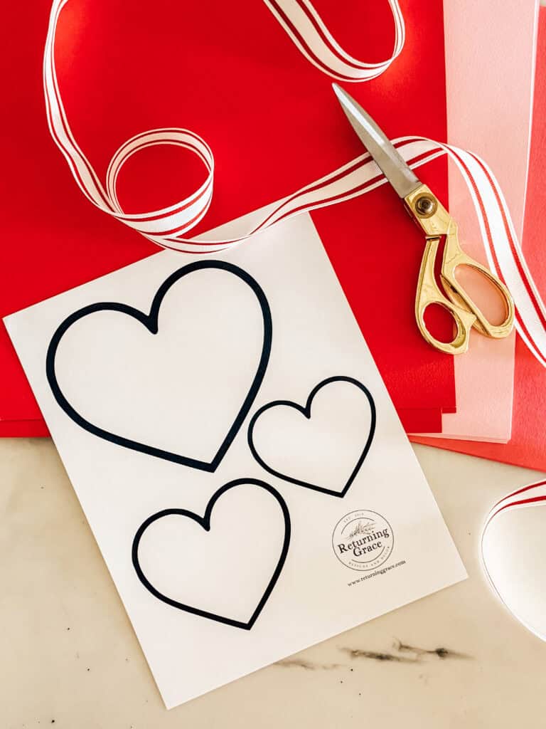 A sheet of paper with three outlined heart shapes sits on a table with red and pink paper sheets, gold scissors, and red-striped white ribbon.