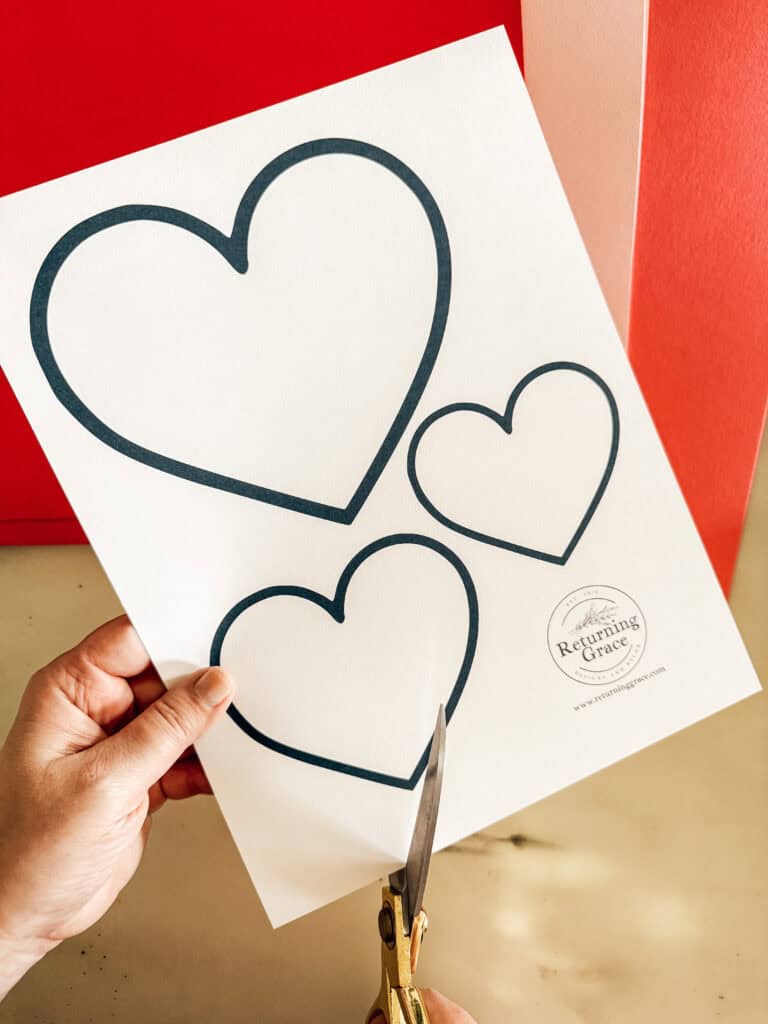 A hand holding scissors is cutting a white sheet of paper with three outlined heart shapes, against a red and pink background. The sheet has a circular logo in the bottom right corner.