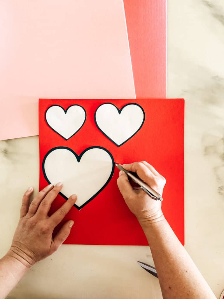 A person traces a large heart shape onto red paper using a craft knife, with two smaller heart shapes above it and pink sheets in the background.