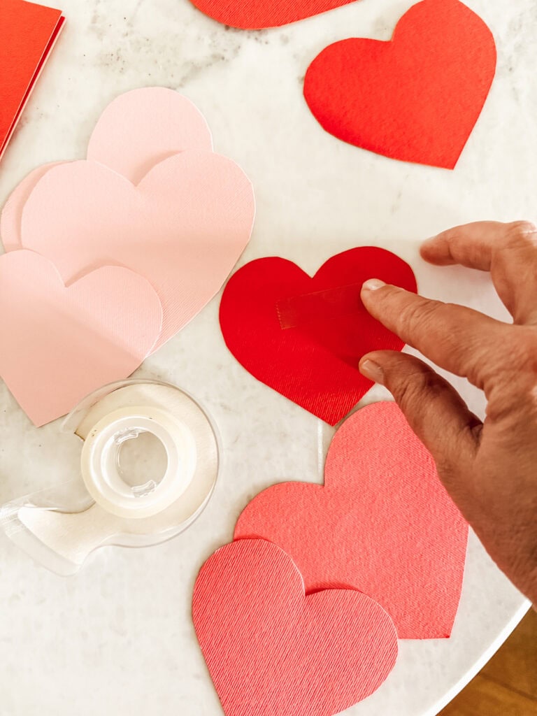 A hand is placing a piece of tape on a red paper heart among several red and pink paper hearts scattered on a white surface. A clear tape dispenser is also visible on the surface.