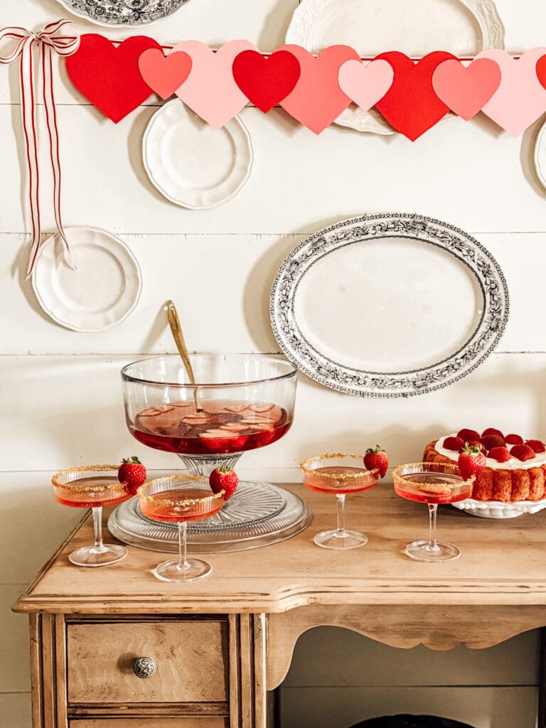 A Valentine’s Day-themed setup with heart garland, vintage plates, a large bowl of punch with fruit, four glasses garnished with strawberries, and a small cake topped with strawberries on a wooden table.