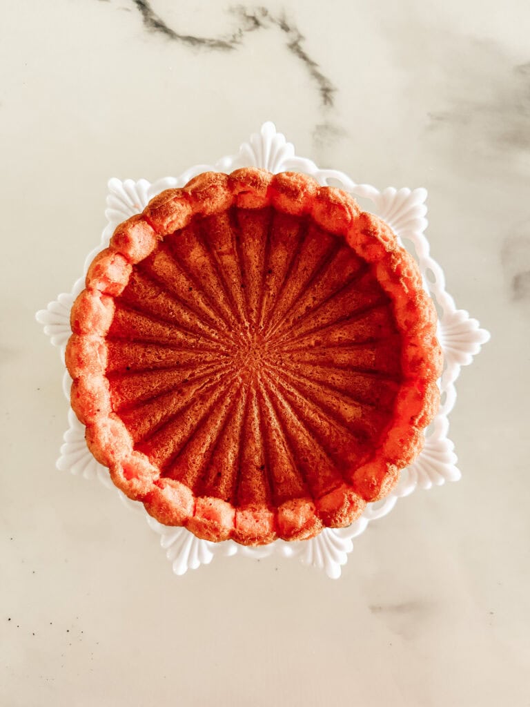 A golden-brown bundt cake with a decorative, fluted pattern sits on a white ornate cake stand against a light marble background, viewed from above.