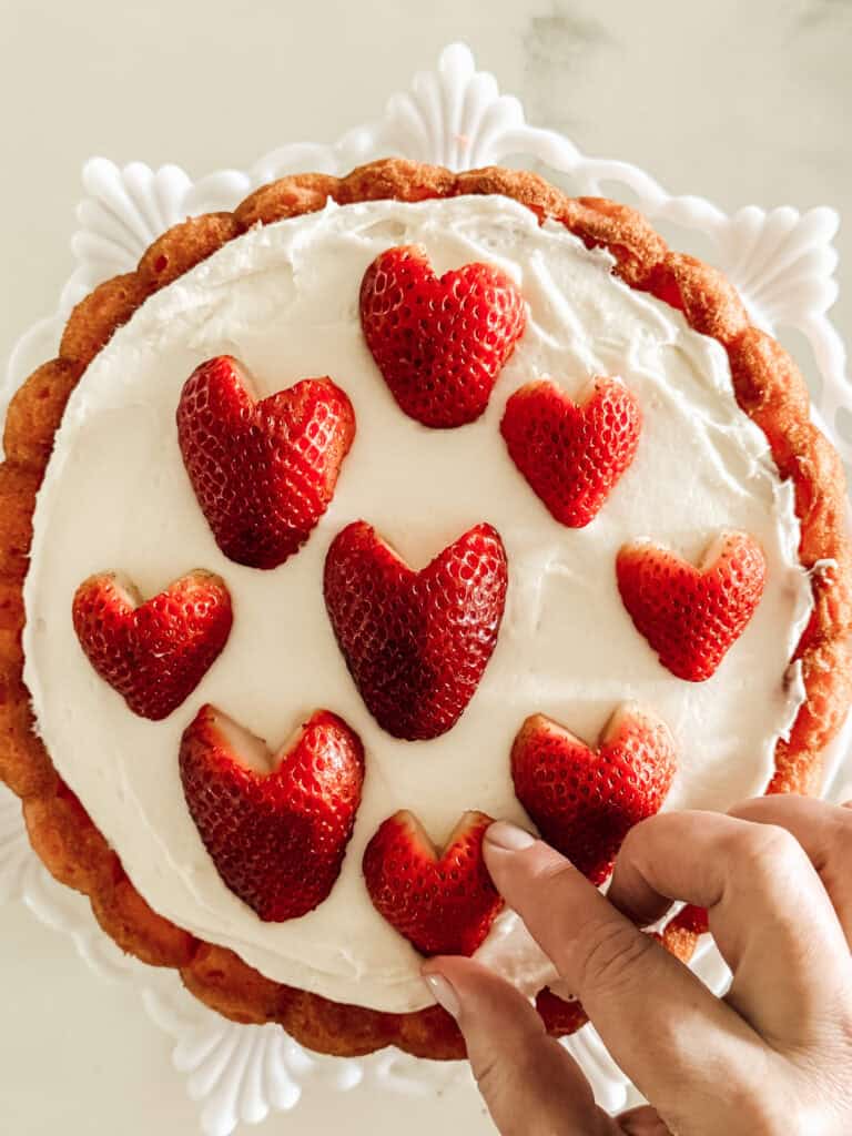 A hand places a heart-shaped strawberry on a cake topped with white frosting and decorated with other heart-shaped strawberries, all arranged in a circle.
