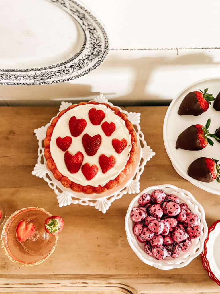 A cake topped with heart-shaped strawberries, chocolate-covered strawberries, a bowl of pink candies, and a glass with a strawberry garnish are arranged on a wooden table.