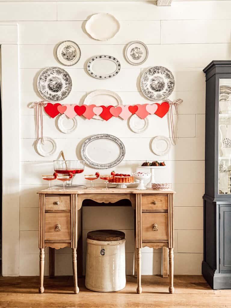 A vintage wooden desk sits against a white paneled wall decorated with displayed plates and a red and pink heart garland. On the desk are bowls of strawberries, desserts, and drinks, creating a festive, cozy atmosphere.