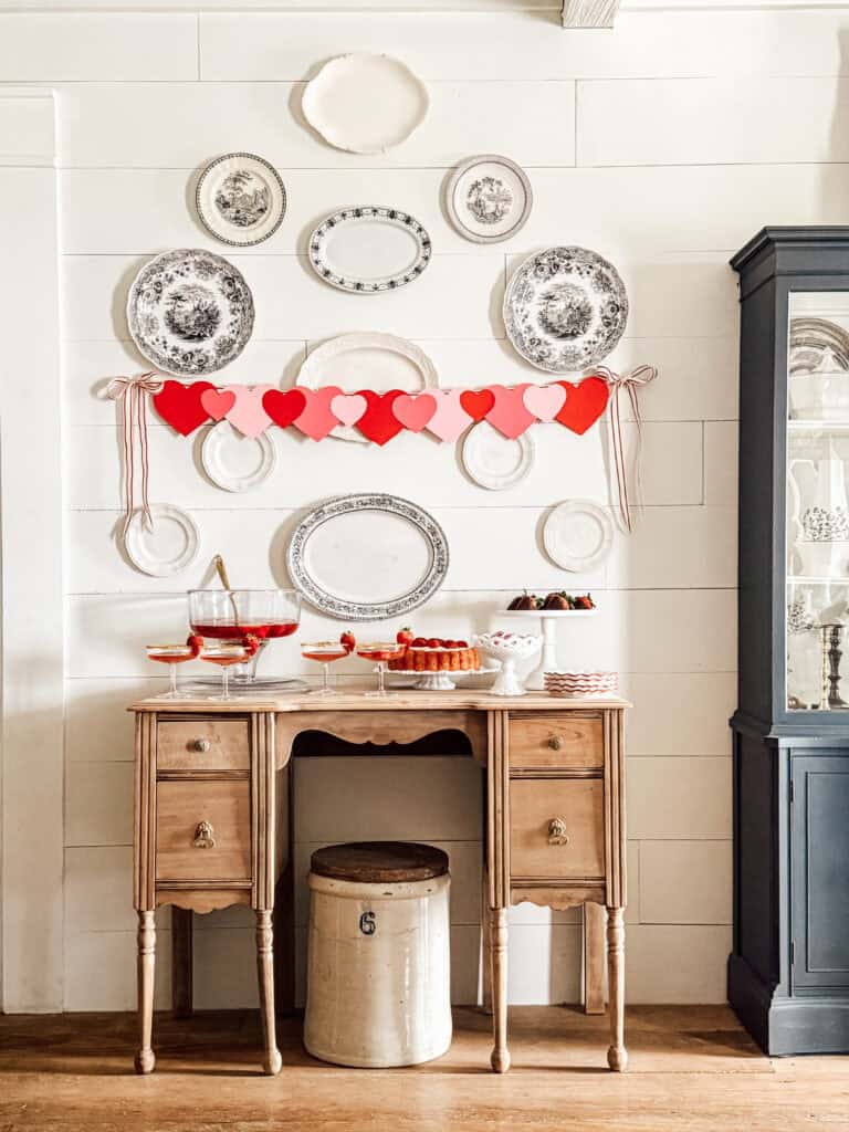 A decorated wooden side table with desserts, punch, and a cake, set against a white shiplap wall with hanging plates and a red and pink heart garland, creating a festive and cozy atmosphere.