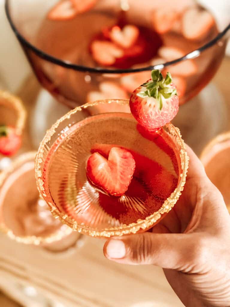 A hand holds a glass of pink punch garnished with a whole strawberry on the rim and a sliced strawberry floating inside. In the background, a punch bowl and more glasses are visible.