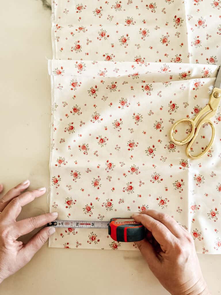 Hands measuring floral fabric with a tape measure next to gold scissors on a white surface. The fabric is cream-colored with small red flowers and green leaves.