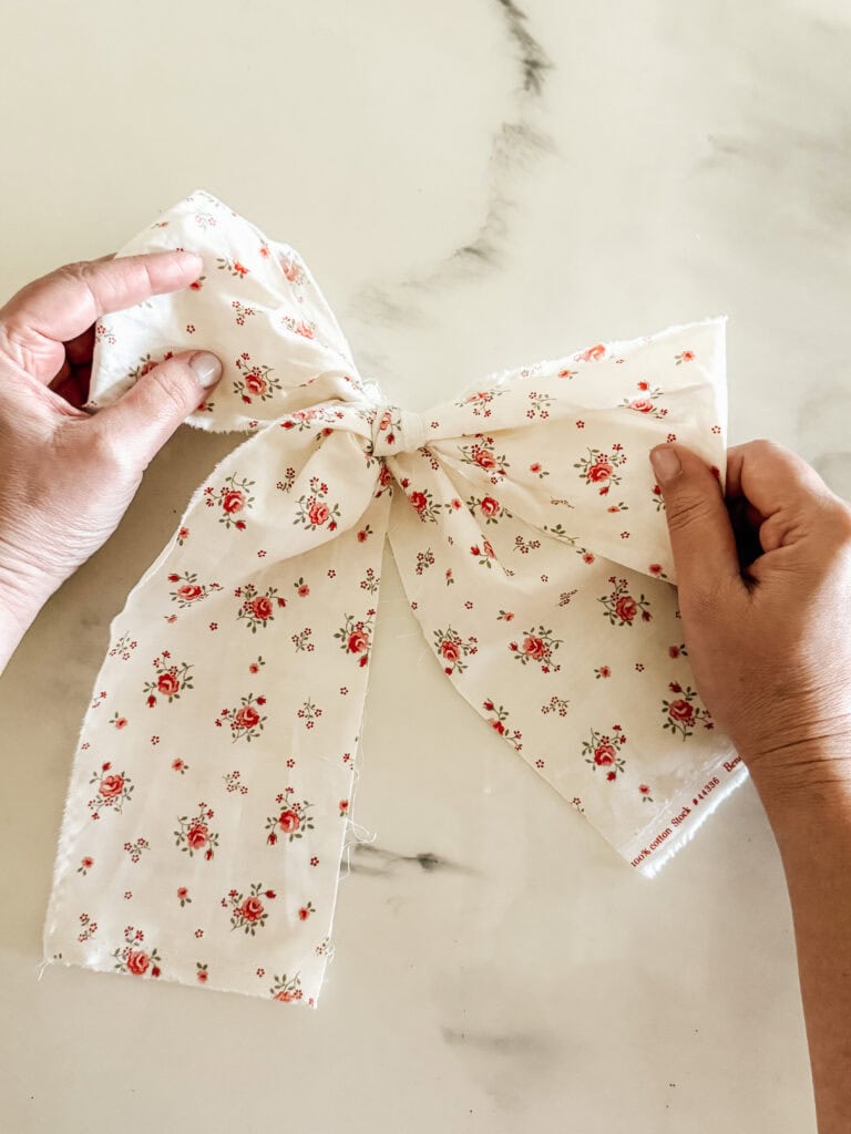 Two hands hold a large, white fabric bow with a red and pink floral pattern, positioned on a light marble surface.