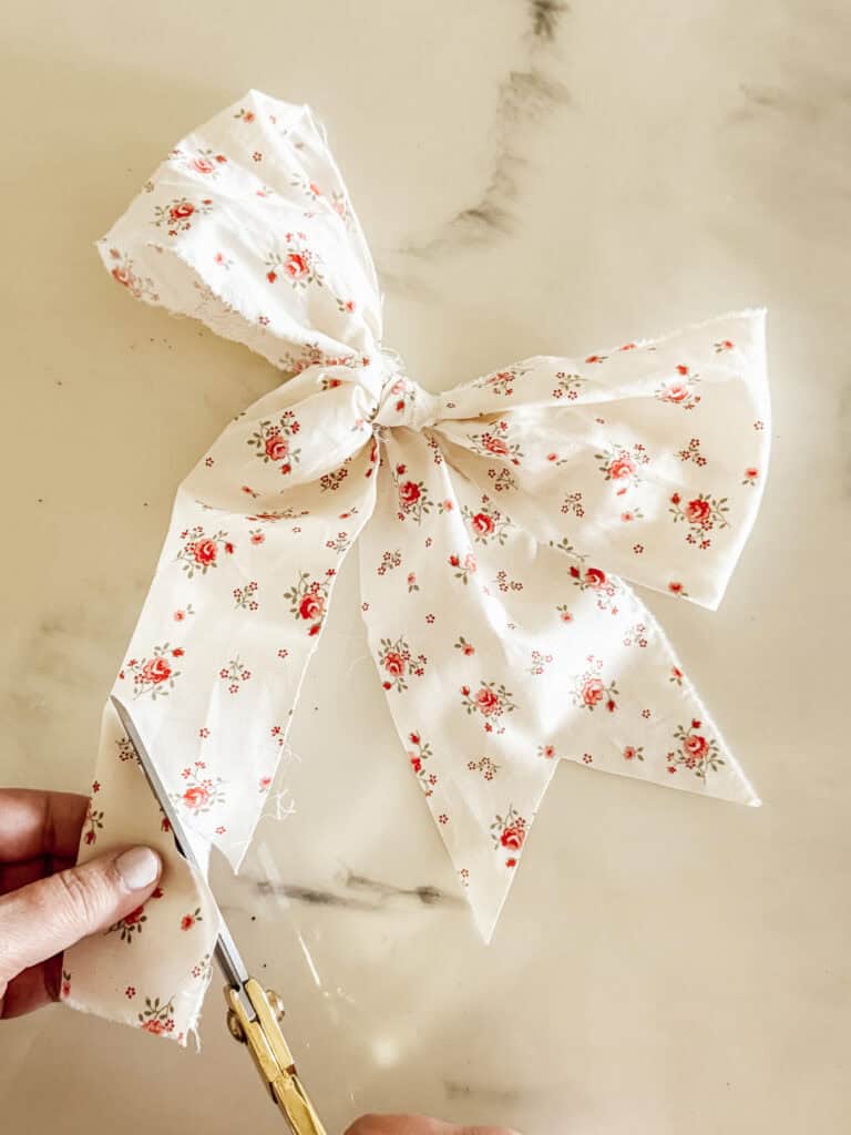 A hand uses gold scissors to trim the end of a large bow made from white fabric with a pink floral pattern, placed on a light-colored marble surface.