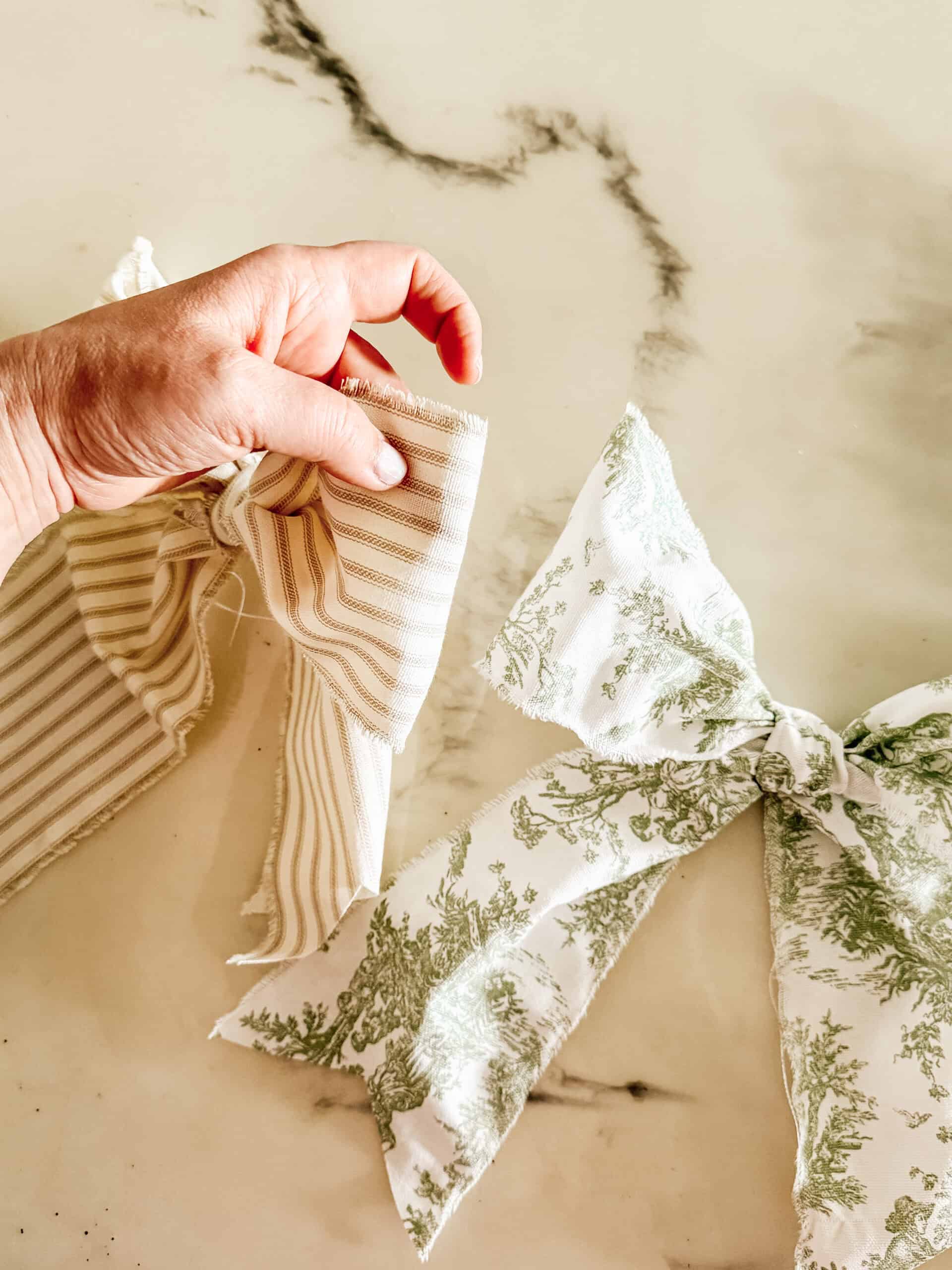 A hand holds a striped fabric bow next to another bow made of white fabric with a green floral pattern, both on a light marble surface.