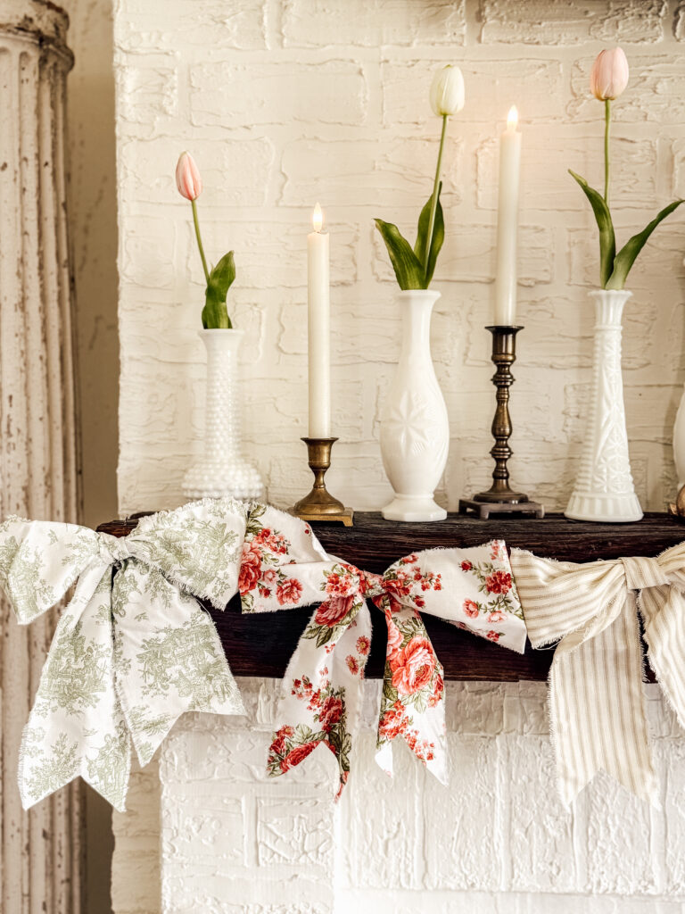 A mantel decorated with white vases holding tulips, brass and white candlesticks with lit candles, and a garland of large fabric bows in floral and striped patterns against a white brick wall.
