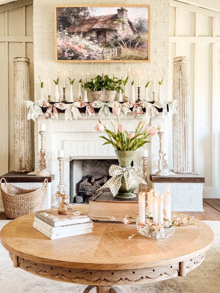 A cozy, light-filled living room with a white brick fireplace decorated with potted plants, ribbons, and candles. A round wooden table holds tulips in a vase, stacked books, and lit candles. A cottage painting hangs above the mantel.