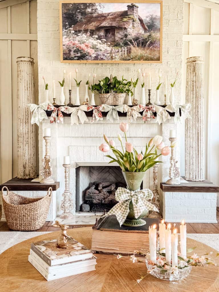 A cozy, vintage-style living room with a white brick fireplace decorated with candles, flower garlands, and plants. A vase of tulips sits on a coffee table with books and lit candles. A cottage painting hangs above the mantel.