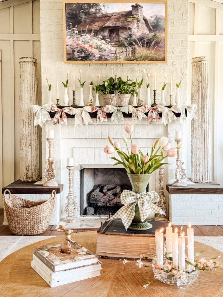 A cozy, vintage-style living room with a white brick fireplace decorated with greenery, candles, and floral garland. A vase of pink tulips, stacked books, and a candle arrangement sit on a wooden coffee table.