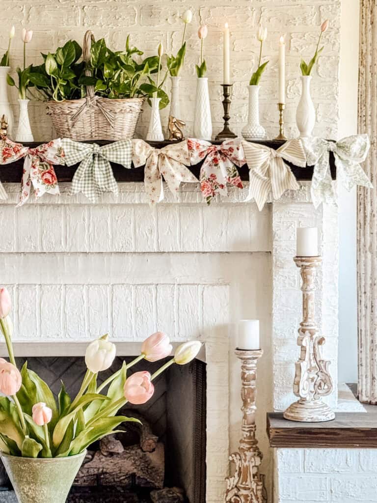 A white brick fireplace decorated with a garland of pastel-patterned bows, white vases holding tulips, potted greenery, and assorted candlesticks. A vase of pink and white tulips sits in the foreground.