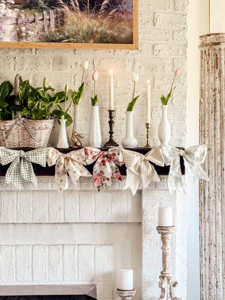 A white brick fireplace mantel decorated with candles, vases holding tulips, a basket of greenery, and large patterned bows in various pastel colors. A framed landscape painting hangs above the mantel.