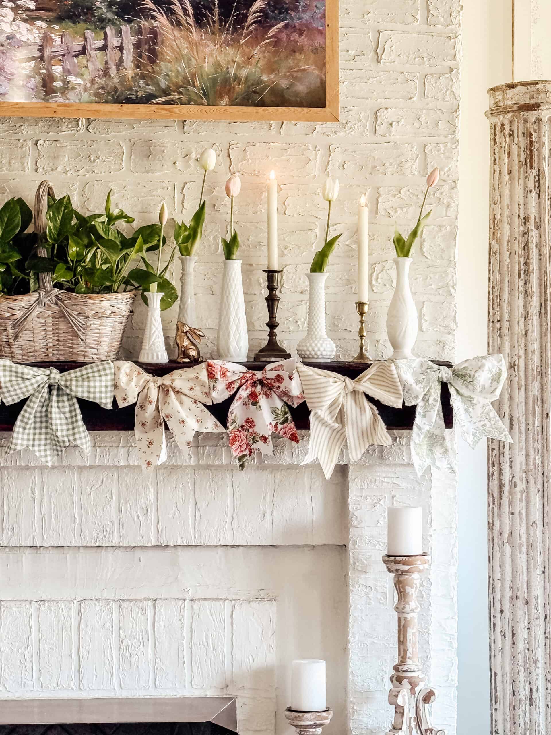 A white brick fireplace mantel decorated with candles, vases holding tulips, a basket of greenery, and large patterned bows in various pastel colors. A framed landscape painting hangs above the mantel.