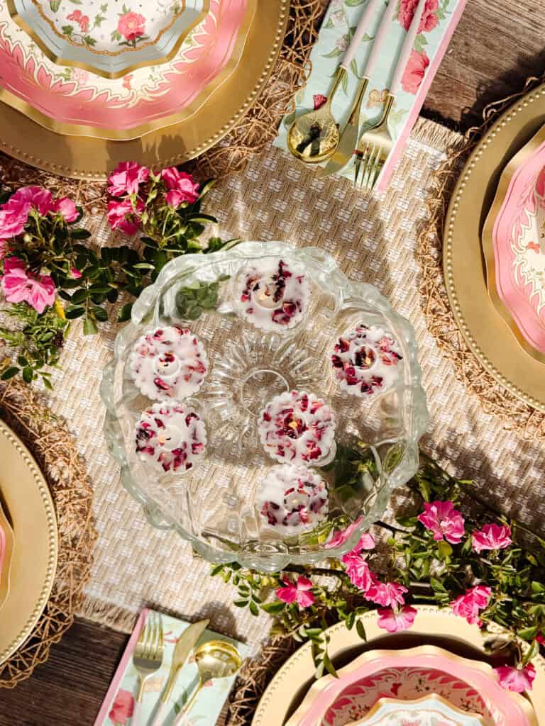 A glass platter with small, white-iced desserts topped with rose petals is set on a woven tablecloth, surrounded by elegant plates, gold utensils, and pink roses as part of a decorative table setting.