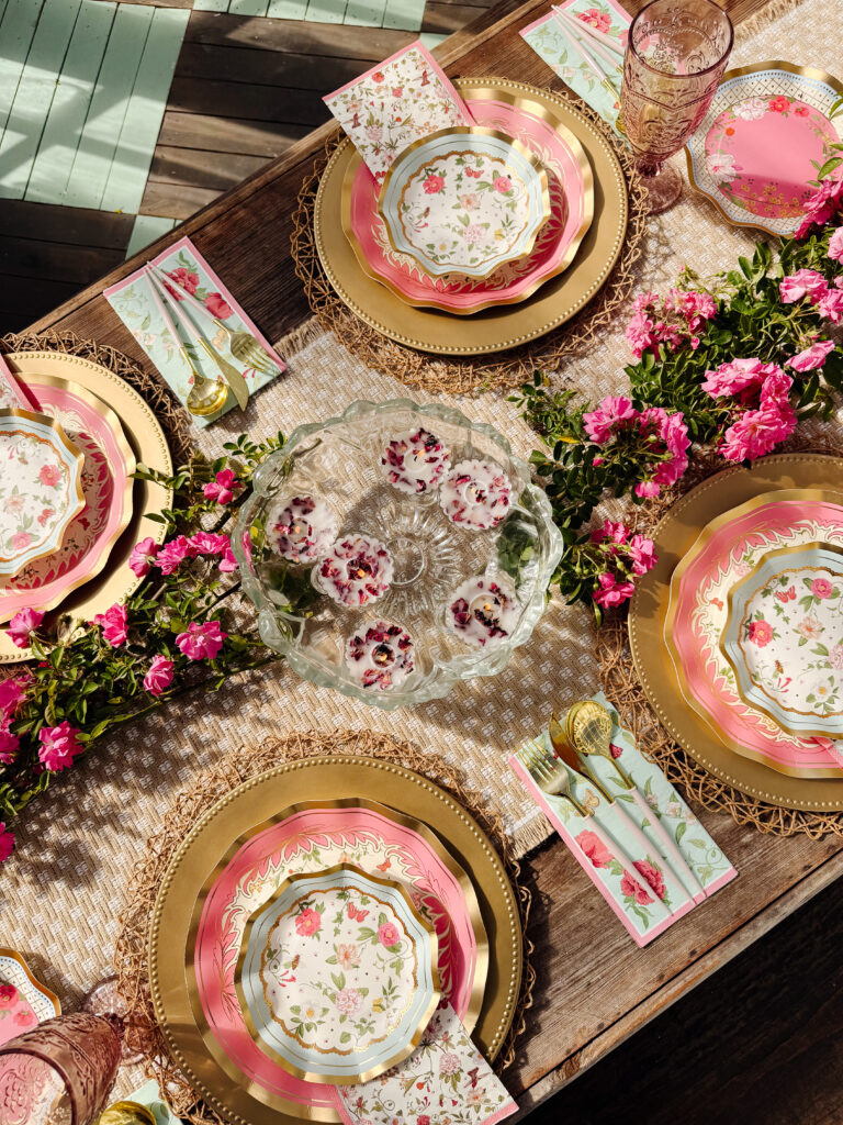 A beautifully set table with floral plates, gold cutlery, pink glasses, and pink flowers arranged along a woven table runner. A glass bowl with floral ice cubes sits at the center, creating a charming, spring-themed setting.