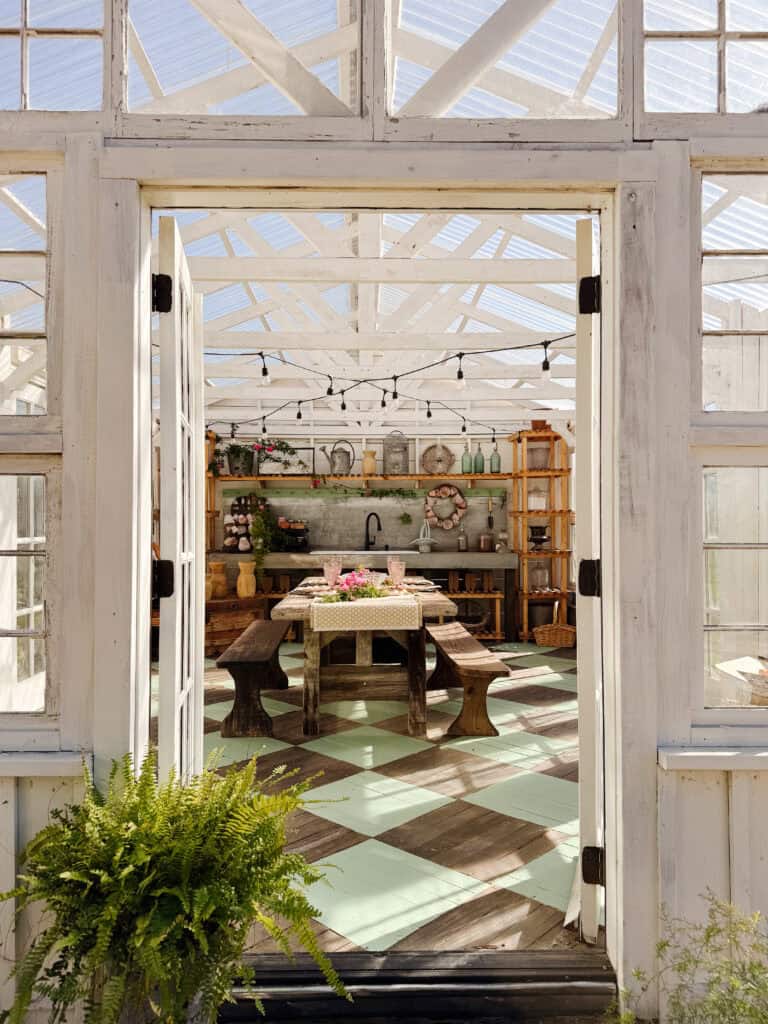 View through open double doors into a bright, rustic greenhouse with a checkered floor, wooden table and benches, potted plants, shelves with gardening supplies, and sunlight streaming through a glass roof.