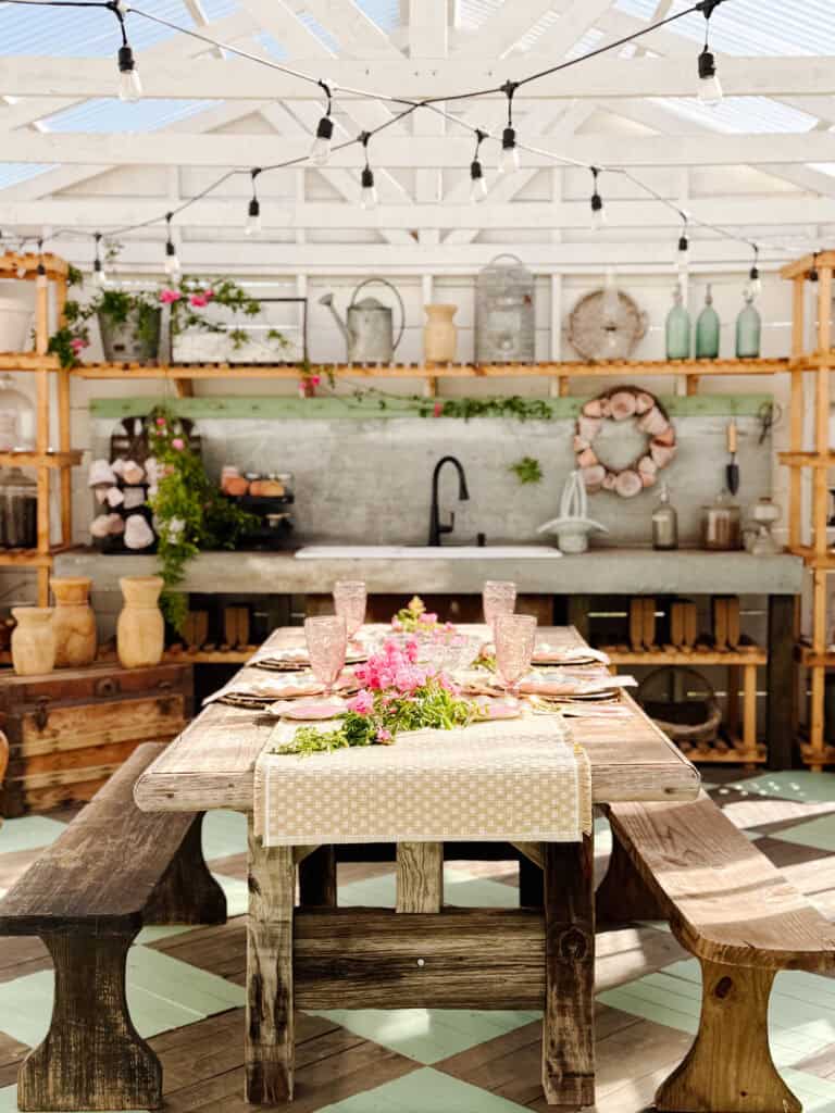 A rustic outdoor dining area with a wooden table set for a meal, pink flowers as a centerpiece, benches, open shelves with pottery, string lights, and plants, beneath a white pergola.