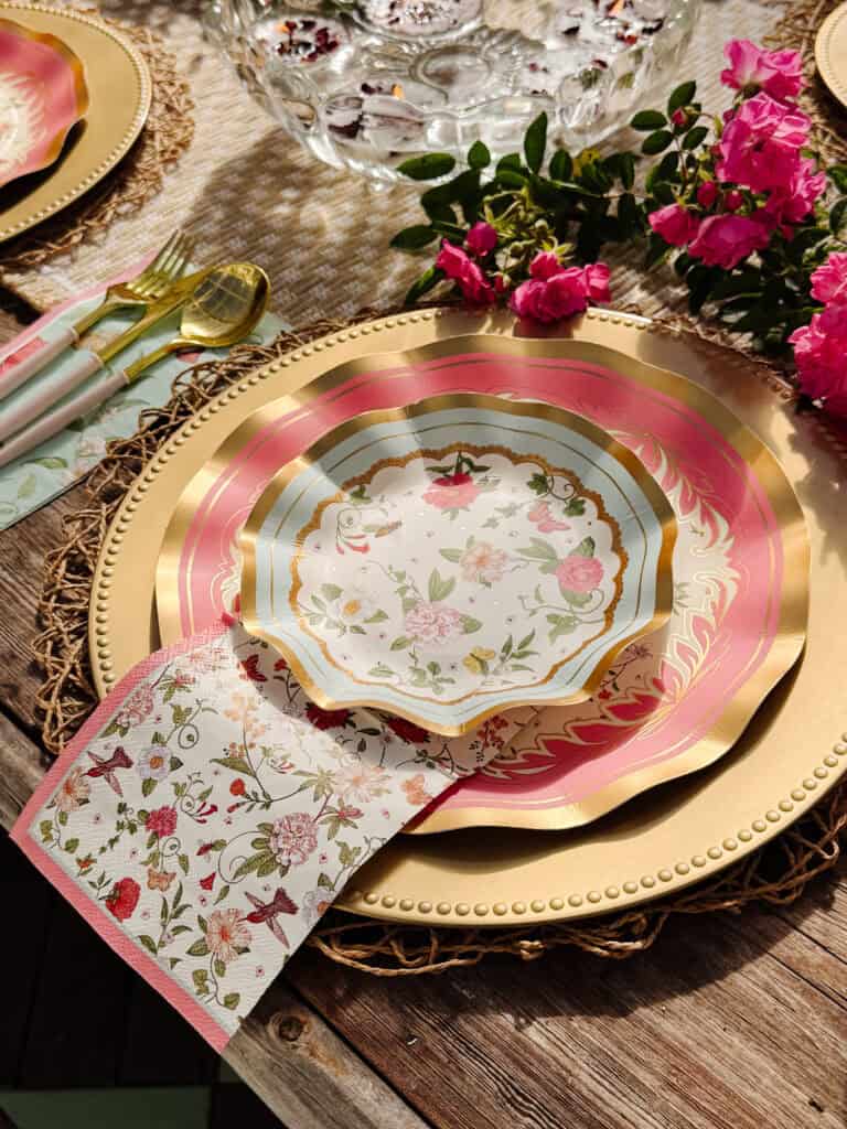 A decorative table setting with gold cutlery, a woven placemat, gold-rimmed floral plates in pink and white, a floral napkin, and pink flowers arranged beside the plates on a rustic wooden table.