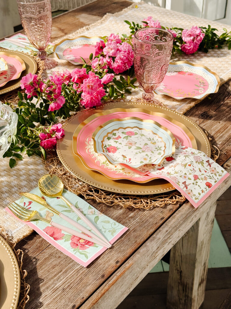 A rustic wooden table set with layered floral plates, gold-rimmed pink glasses, gold and pastel cutlery on floral napkins, and bright pink flowers as a centerpiece. The setting has a romantic, spring-inspired theme.