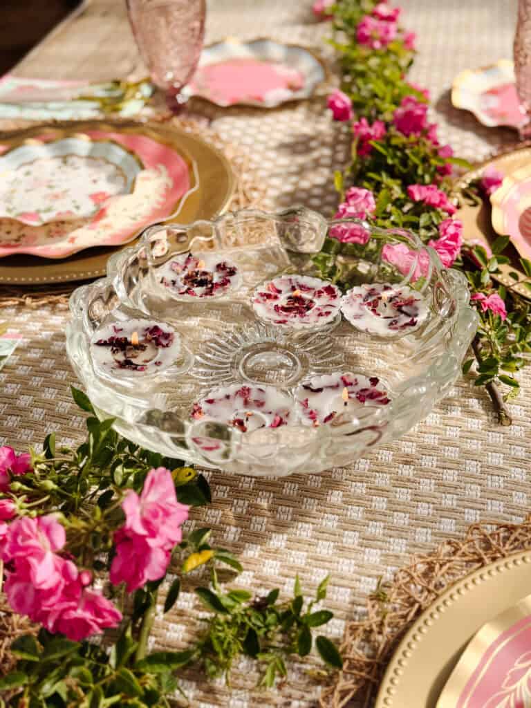 A glass punch bowl holds floating white candles with flower petals. The bowl sits on an elegant table set with pink and gold plates, pink glasses, and a floral garland of vibrant pink flowers.