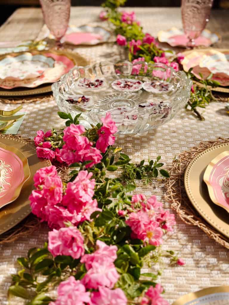 A decorated table with gold-accented plates, pink glasses, a clear glass bowl with floating flowers, and a centerpiece of vibrant pink flowers and greenery running down the middle.