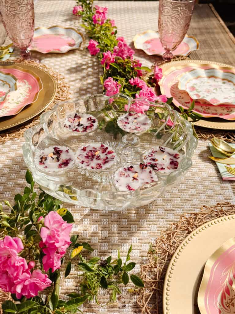 A decorative outdoor table setting features pink flowers, layered plates with gold accents, pink glasses, and a glass bowl with floating candles containing flower petals. Bright sunlight highlights the elegant, festive arrangement.