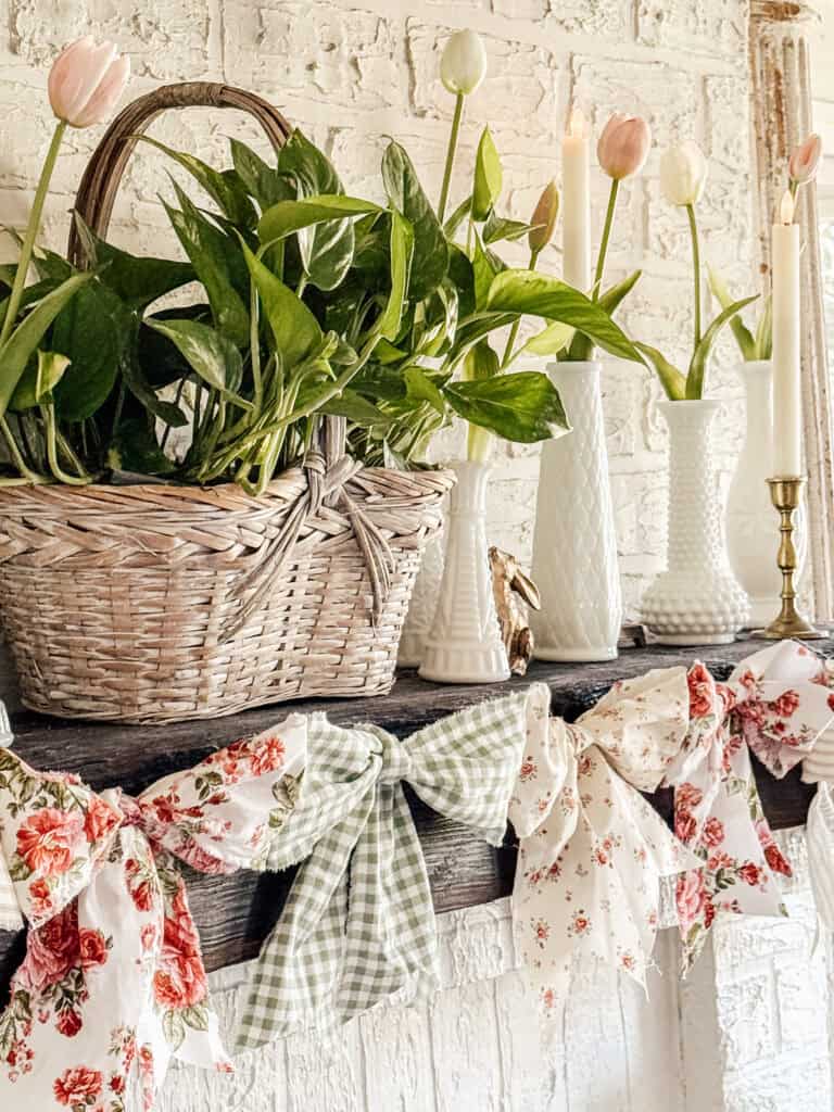 A rustic mantel decorated with a woven basket of green plants, white vases holding pastel tulips, and large floral and gingham bows draped along the edge against a white brick background.