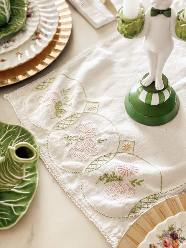 A close-up of a table setting with a white embroidered table runner featuring pink and green floral designs, green leaf-shaped plates, gold-rimmed dishes, and a white rabbit candlestick holder.