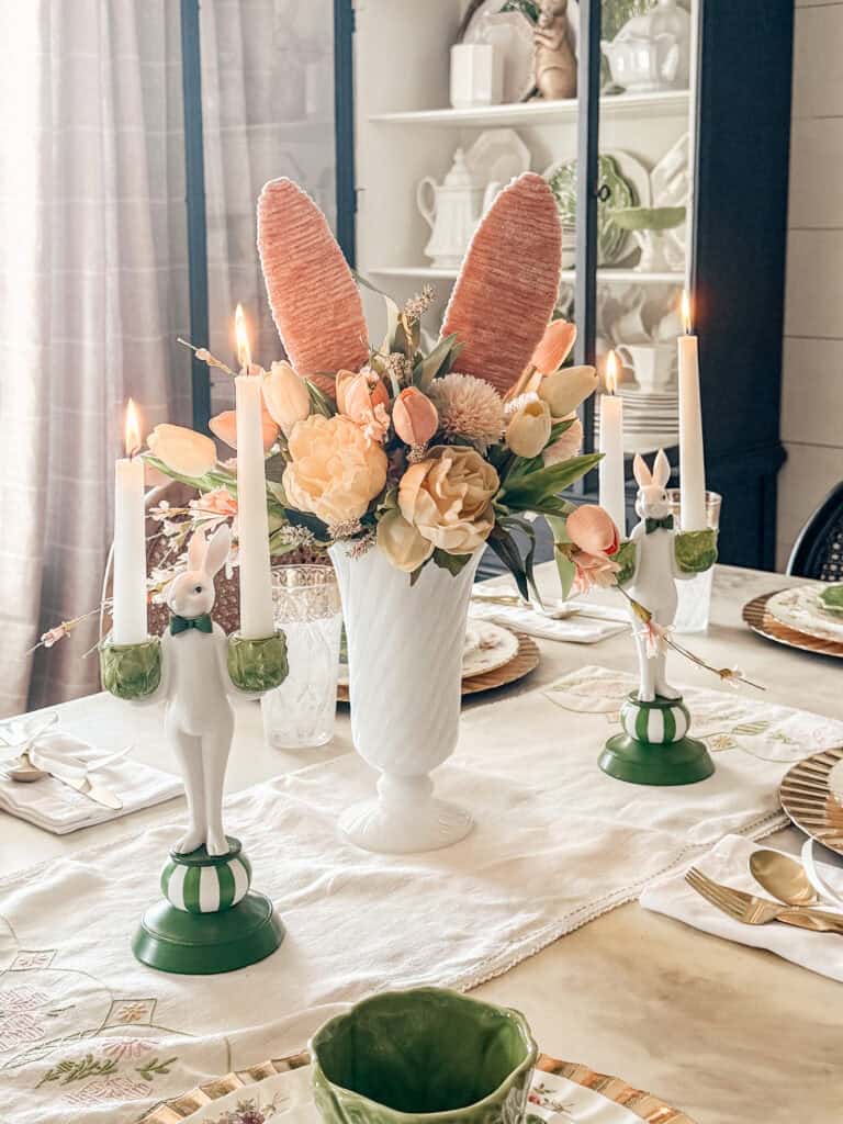A dining table is set with floral centerpiece featuring bunny ears, two lit bunny-shaped candlesticks, and elegant tableware. The scene is soft and pastel, evoking a spring or Easter theme.