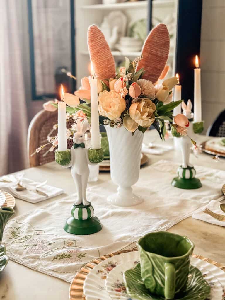 A spring-themed table setting with green and white rabbit candlestick holders, tall white candles, a white vase of flowers with large bunny ears, green leaf-shaped teacups, and gold-edged plates on a cream tablecloth.