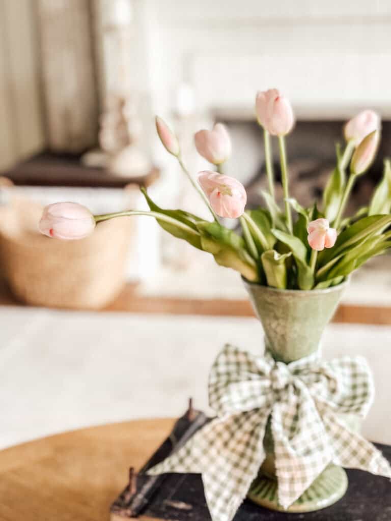 A green vase with light pink tulips sits on a table, decorated with a checkered green and white ribbon tied in a bow. The background is softly blurred, showing home decor elements.