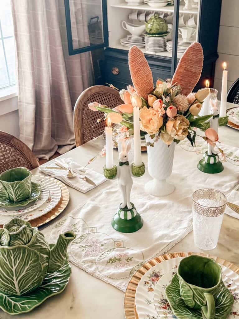 Elegant spring table setting with cabbage-shaped teapot and cups, floral plates, lit candles in bunny holders, and a centerpiece of peach and cream flowers with bunny ear decorations. Light streams in through a nearby window.