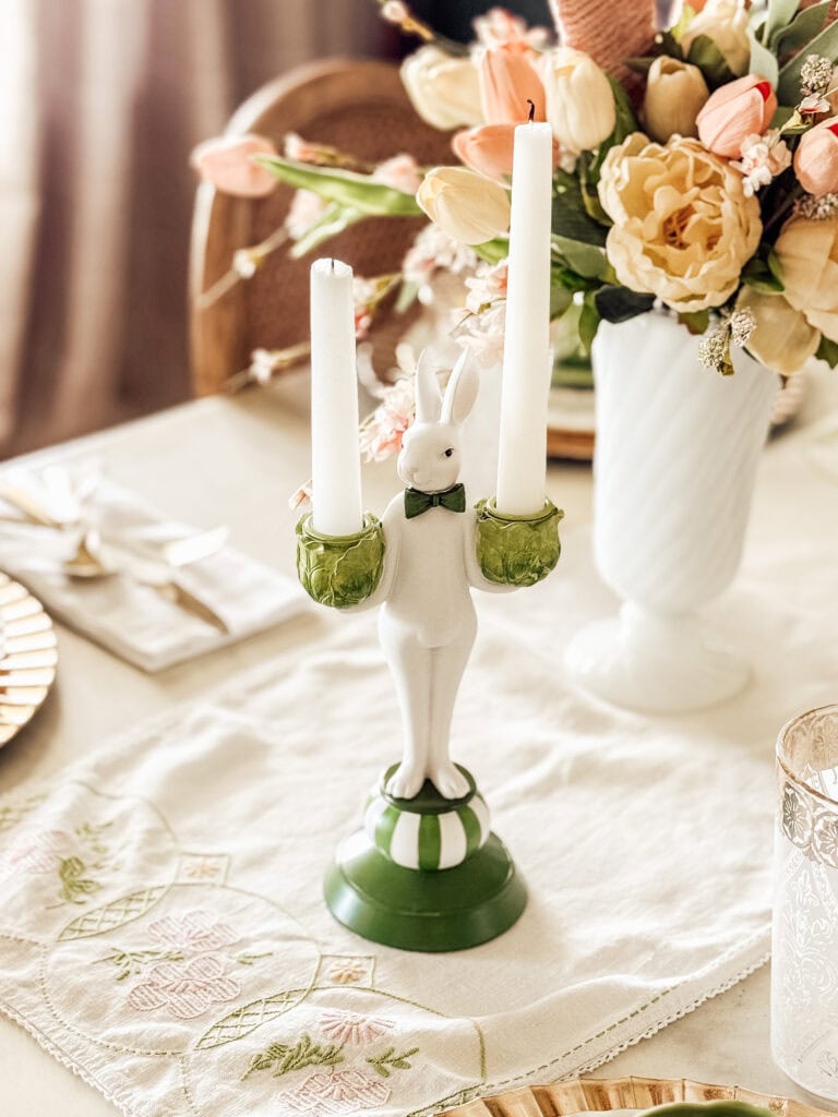A ceramic bunny candle holder with a green bow tie holds two white candles on a decorated table with a floral arrangement and embroidered tablecloth.