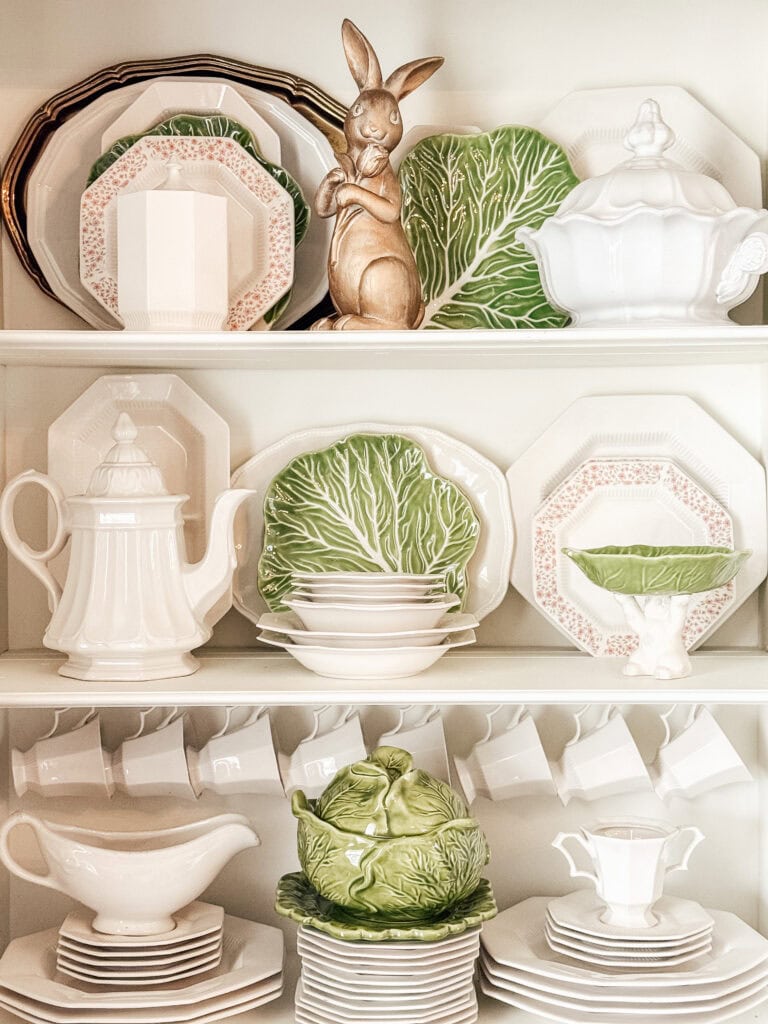 A kitchen shelf displays white dishes, teacups, and serving pieces, accented with green cabbage-shaped plates and a bowl. A gold rabbit figurine stands among the items. The arrangement is neat and decorative.
