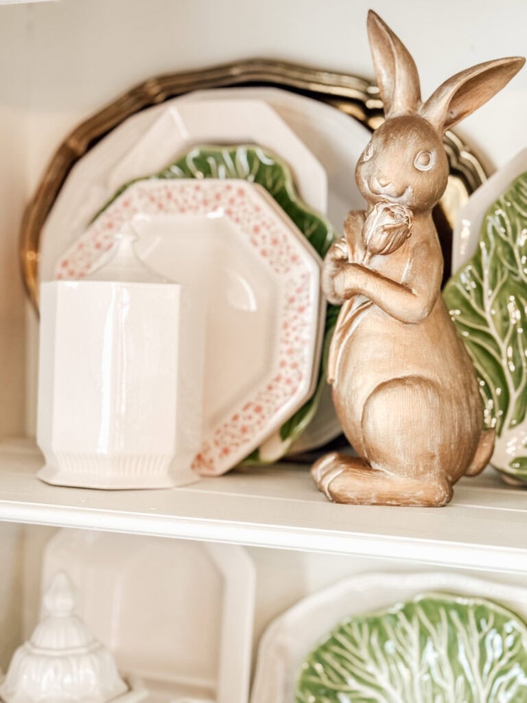 A gold bunny figurine holding tulips is displayed on a shelf next to white, floral, and green leaf-shaped plates, creating a decorative and spring-themed arrangement.