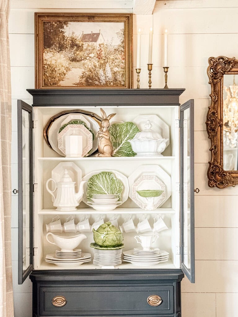 A black cabinet with glass doors displays white and green dishware, including cabbage-shaped plates, teacups, and a rabbit figurine. On top are a framed painting and three unlit candlesticks next to a gold-framed mirror.