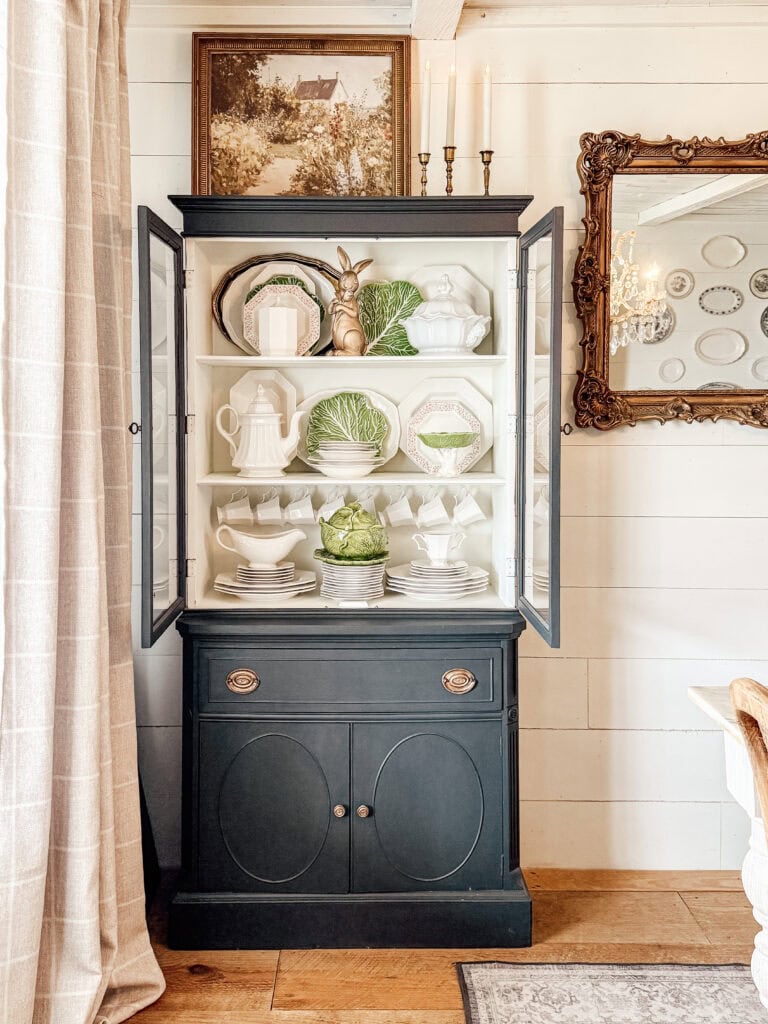 A black china cabinet with glass doors displays white and green dishes, including leaf-shaped plates and a gold bunny figure. The cabinet sits against a white shiplap wall, with framed art and a mirror nearby.