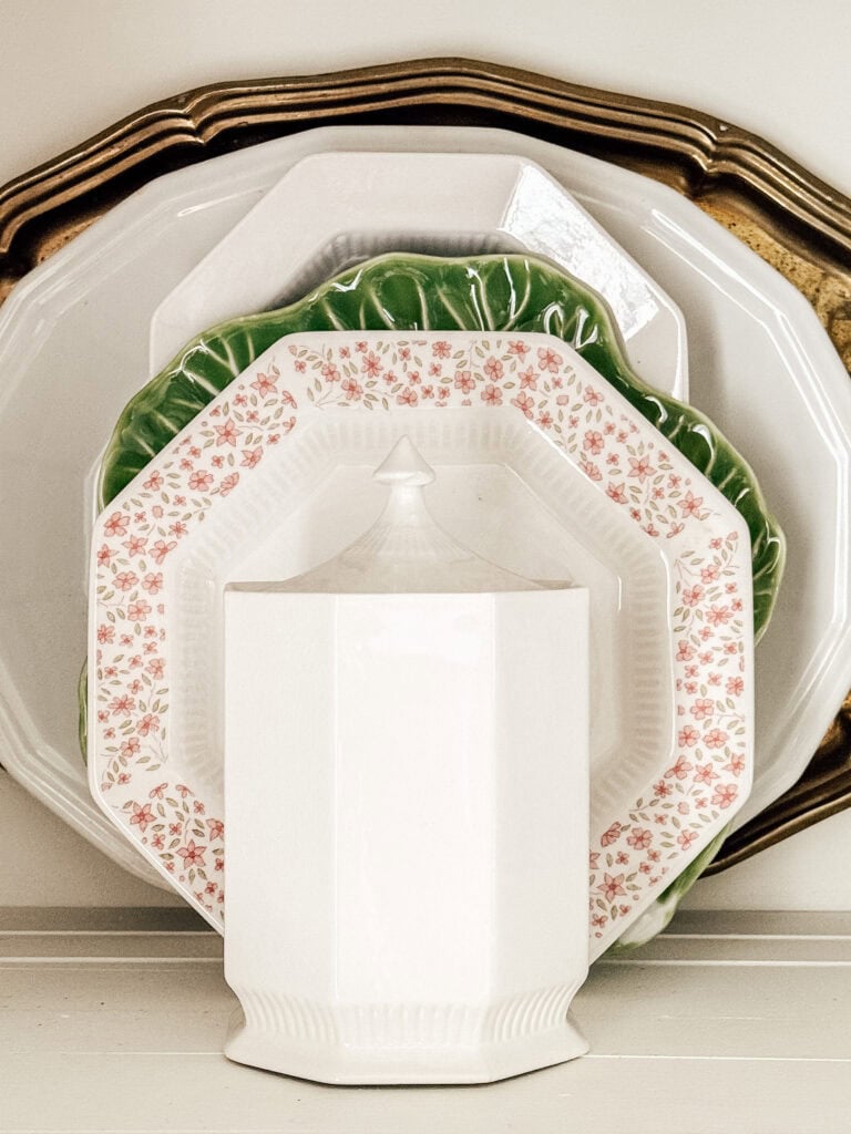 A stack of assorted plates, including a gold-rimmed plate, a green leaf-shaped plate, and an octagonal plate with a pink floral pattern, stands behind a white geometric teapot on a shelf.